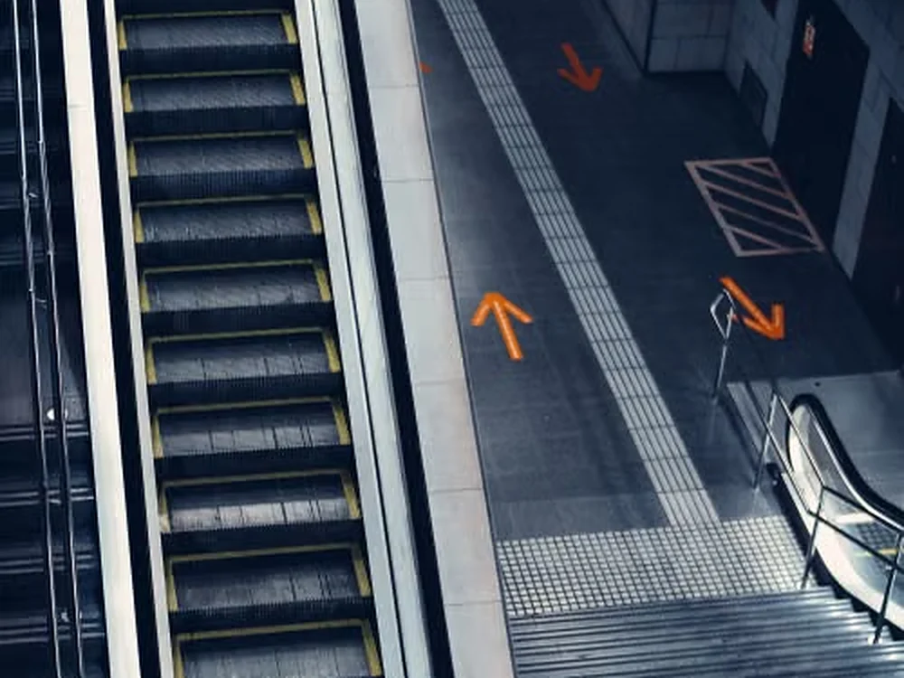 Escalator steps with yellow edge markings beside floor direction arrows and tactile paving at a landing